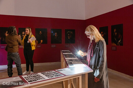 Drucke einer Fotostrecke sind auf einem sehr langen, schmalen Tisch zu sehen und werden von einer Besucherin mit einer Handytaschenlampe angeleuchtet. Im Hintergrund unterhalten sich zwei Menschen vor Portraitreihen an der Wand.