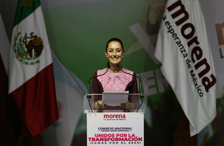Claudia Sheinbaum steht an einem Redner*innenpult vor zwei Fahnen, einmal der von Mexiko und einmal der ihrer Partei.