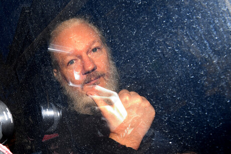 Julian Assange durch eine Scheibe hindurch aufgenommen. Es ist noch ein Kameraobjekt einer anderen Kamera zu sehen, die Reflektion von Blitzlicht erscheint auf der Scheibe.