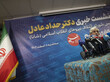 Ein iranischer Politiker steht vor mehreren Mikrofonen, neben ihm eine iranische Flagge.