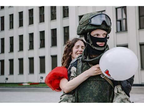 Eine junge Frau in einem roten Kleid, die einen weißen Luftballon in einer Hand hält, umarmt weinend einen Soldaten, der behelmt und in Tarnkleidung in eine andere Richtung schaut.