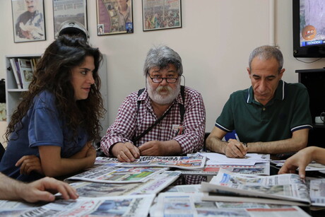 Drei Personen sitzen an einem Tisch, auf dem Zeitungen liegen.