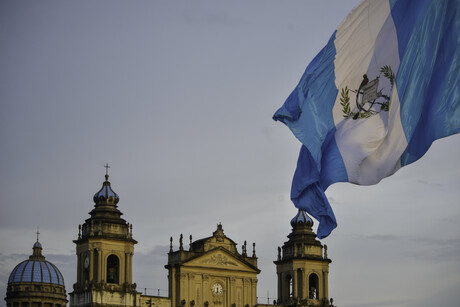 Guatemala-Stadt; auf der linken Bildseite ist ein herrschaftliches Gebäude zu sehen, auf der rechten Seite weht die Nationalflagge (vertiakl blau-weiß gestreift)