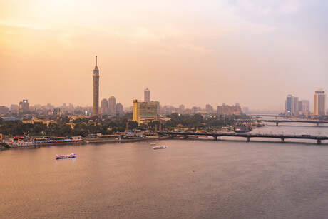 Vom Nil aus kann man über Kairo mit seinen Hochhäusern und Parks schauen; auf dem Fluss liegen einige Schiffe und rechts im Bild sind zudem noch drei Brücken zu sehen