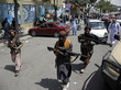 Drei bewaffnete Männer in traditioneller afghanischer Kleidung gehen über eine Straße. Im Hintergrund weitere Männer und mitten auf der Straße abgestellte Autos