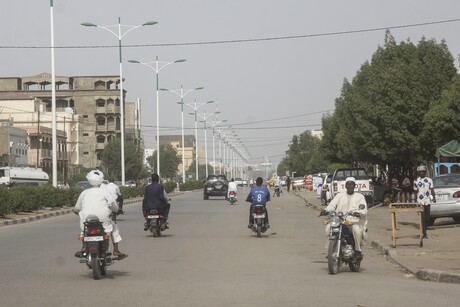 Auf einer Straße fahren Motorräder und Autos; links von der Straße sind außerdem Gebäude und Straßenlaternen und rechts Bäume zu sehen 