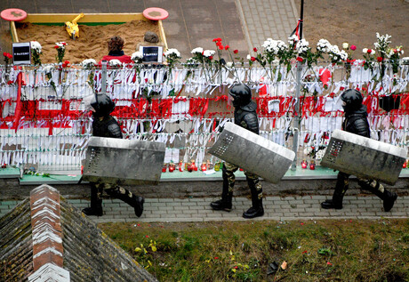 Drei Belarusische Einsatzkräfte laufen an einem Zaun vorbei, der mit weißen und roten Bändern sowie Rosen geschmückt wurde