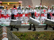 Drei Belarusische Einsatzkräfte laufen an einem Zaun vorbei, der mit weißen und roten Bändern sowie Rosen geschmückt wurde