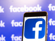 Auf einem Handy ist ein weißes f, das Logo von Facebook, zu sehen. Dahinter steht auf einer Wand mehrmals in weißer Schrift auf blauem Grund Facebook.