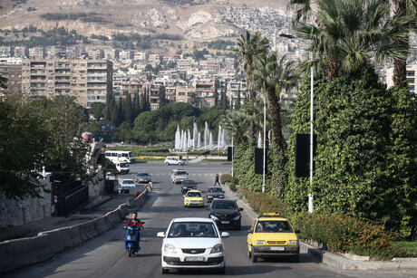 Auf einer Straße von Damaskus fahren Autos und Motorroller; am linken und rechten Starßenrand stehen Bäume. Im Hintergrund ist ein Kreisverkehr mit einem großen Springbrunnen zu sehen, hinter dem sich helle Häuser über weite Berge erstrecken.