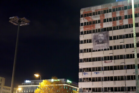 Vor einem dunklen Abendhimmel ist ein Gebäude zu sehen, an dessen Außenfront ein Banner mit dem Gesicht von Khaled Drareni hängt