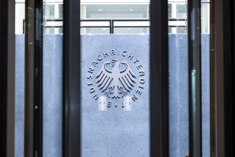 Auf dem Bild ist eine Glastür abgebilet, durch die Tür sieht man das metallene Logo des Bundesnachrichtendienstes. 