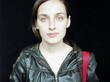 Portraitfoto von Jelena Kostjutschenko vor schwarzem Hintergrund. Sie trägt eine schwarze Lederjacke und hat einen markanten, roten Henkel über der Schulter hängen, welcher aufgrund der roten Farbe stark auffällt.