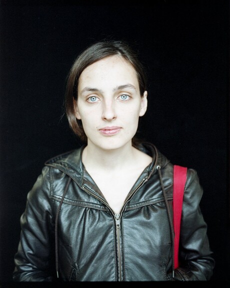 Portraitfoto von Jelena Kostjutschenko vor schwarzem Hintergrund. Sie trägt eine schwarze Lederjacke und hat einen markanten, roten Henkel über der Schulter hängen, welcher aufgrund der roten Farbe stark auffällt.