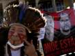 Ein Indigener mit Feder-Kopfschmuck bemalt sein Gesicht, hinter ihm ein Protestplakat mit zwei Gesichtern und der Aufschrift "Wo sind?" auf Portugiesisch.