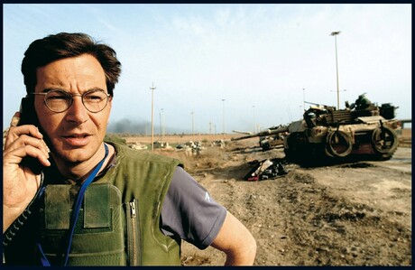 Ein Mann mit Brille und schussicherer Weste telefoniert. Im Hintergrund sind mehrere Panzer auf einer sandigen Straße zu sehen.