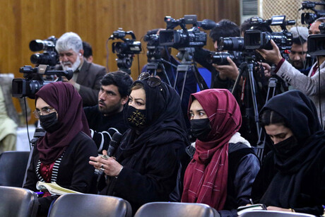 Mehrere Journalist*innen stehen und sitzen in einer Presekonferenz. Die sitzenden Frauen tragen Kopftücher sowie medizinische Masken, die dahinter sitzenden und stehenden Männer allesamt nicht.