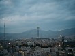 Teheran erstreckt sich in der Dämmerung vor einem Gebirgszug; ein Fernsehturm hebt sich im Mittelpunkt des Bildes von den umliegenden, dezent beleuchteten Gebäuden ab