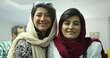 Auf dem Bild sind zwei lächelnde Frauen abgebildet. Links ist Nilufar Hamedi, Rechts ist Elahe Mohammadi. Nilufar ist ein kleines Stück größer und hat etwas hellere braune Haare als Elahe. Sie trägt eine Schwarz-Weiß gemusterte Bluse und hat eine Sonnenbrille auf dem Kopf. Elahe trägt ein weißes Hemd mit schmalen schwarzen Streifen. Beide Frauen tragen eine Schaila, Nilufars ist Creme-Weiß, Elahes ist Purpur-rot. 