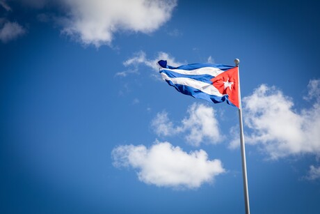 Vor einem blauen Himmel mit weißen Wolken weht die kubanische Flagge: blau-weiße Streifen und ein rotes Dreiek auf der rechten Seite, in dessen Mitte ein weißer Stern zu sehen ist
