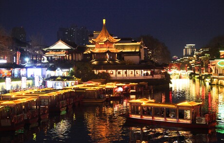 Die chinesische Stadt Nanjing bei Nacht: auf dem Wasser liegen erleuchteten Boote und dahinter sind ebenfalls zahlreiche erleuchtete Gebäude zu sehen