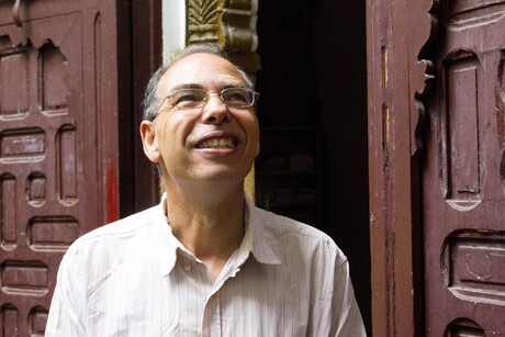 Maati Monjib steht vor einem Hausgang und lächelt in den Himmel; er hat kurze graue Haare, trägt eine Brille und ein helles Hemd