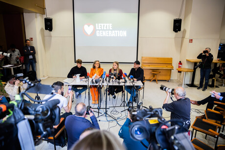 Vier Personen sitzen an einem Tisch, viele Mikrofone und Kameras auf sie gerichtet, hinter ihnen ist der Schriftzug "Letzte Generation" an die Wand projiziert