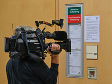 Ein Journalist steht mit einer Kamera auf der Schulter vor einem geschlossenen Gerichtssaal