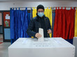 Ein Mann steckt einen Umschlag in eine Wahlurne; hinter ihm sind Wahlkabinen in den Farben der rumänischen Nationalflagge zu sehen