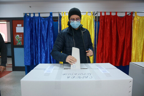 Ein Mann steckt einen Umschlag in eine Wahlurne; hinter ihm sind Wahlkabinen in den Farben der rumänischen Nationalflagge zu sehen
