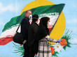 Eine Frau mit Kopftuch und zwei Männer, alle mit Mundschutz, laufen vor einer Wandmalerei vorbei, die eine iranische Flagge zeigt.
