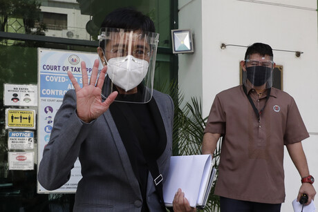 Maria Ressa steht mit Maske und Gesichtsschild winkend vor einem Gebäude; rechts hinter ihr ist ein weiterer Mann zu sehen