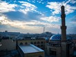 Eine Stadt erstreckt sich vor einem Himmel mit untergehender Sonne; im rechten Vordergrund ist noch die Kuppel und der Turm einer Moschee zu sehen