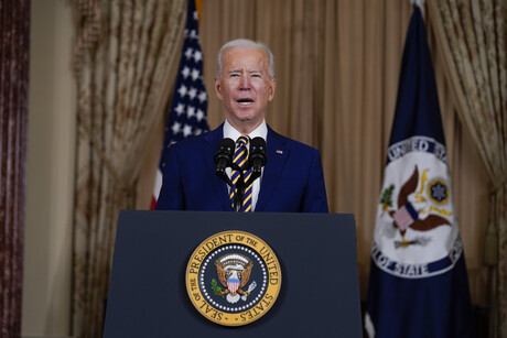 Joe Biden steht an einem Rednerpult, an dessen Frontseite das Symbol der USA zu sehen ist: der Weißkopfadler. Im Hintergrund ist die US-amerikanische Flagge sowie eine weitere Flagge mit dem Weißkopfseeadler zu sehen.
