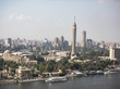 Hinter dem Nil stehen, umgeben von Bäumen und Palmen, unzählige Gebäude; ein hoher Turm auf der rechten Bildseite sticht besonders hervor