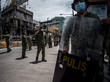 Philippinische Polizisten stehen über eine Straße versteut mit Masken dar, ein Polizist im rechten Vordergrund hält auch ein Schild vor dem Körper; im Hintergrund sind graue Häuser zu sehen